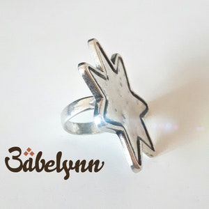 May include: A silver ring with a star-shaped design. The star is textured and has a slightly raised edge. The ring is a simple band.