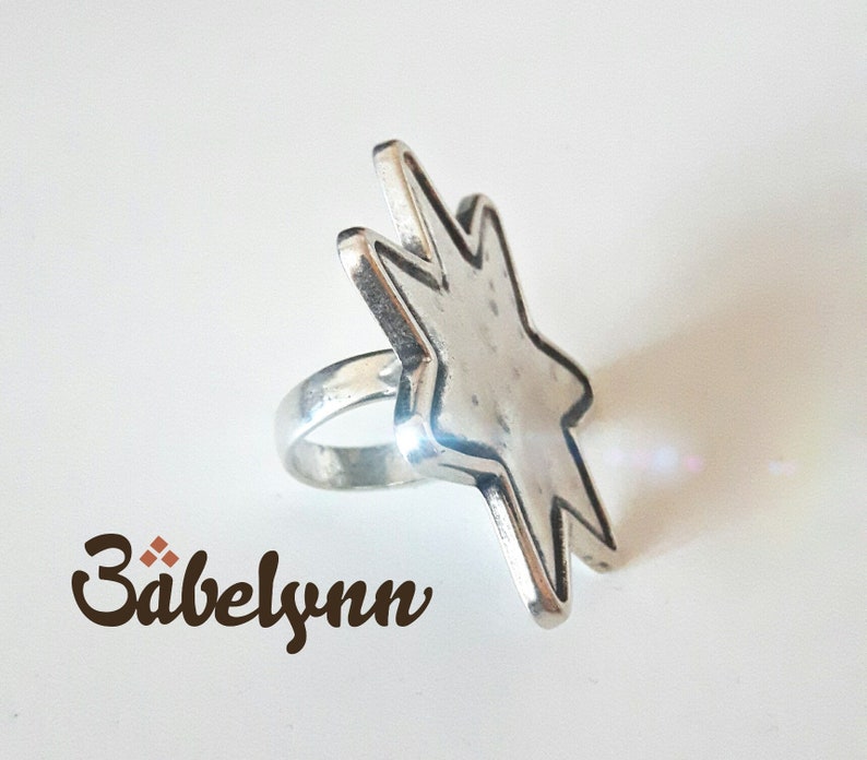 May include: A silver ring with a star-shaped design. The star is textured and has a slightly raised edge. The ring is a simple band.