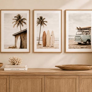 Peut inclure: Trois cadres muraux représentant des scènes de plage dans des tons sépia et doux. Le premier montre une planche de surf contre une cabane en bois, le second trois planches sur le sable avec un palmier, et le troisième un van vintage sur la plage.