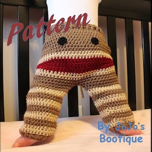 Puede incluir: Pantalones de bebé tejidos a crochet con un diseño de cara de mono. Los pantalones son de rayas marrones, beige y rojas. El patrón se titula "Pattern by JoJo's Boutique".