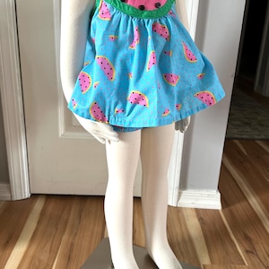 May include: A blue and pink sleeveless dress with a watermelon print. The dress has a green trim and buttons down the front.