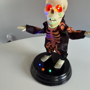 May include: A Halloween-themed animated skeleton figure. The skeleton has glowing red eyes and is wearing a black outfit with an orange skeleton design. It stands on a black base with colored lights.
