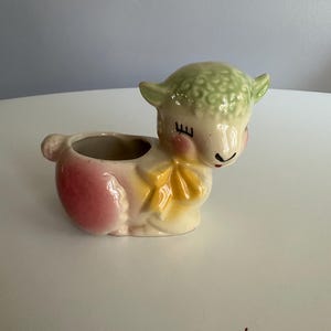 May include: A ceramic planter shaped like a lamb with a light green textured head, pink cheeks, and a yellow bow around its neck. The body features a hollow opening for plants, with a pink-tinted rear section and a small tail on the back side.