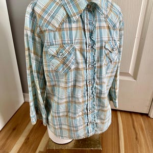 May include: A light blue, tan, and white plaid button-up shirt with long sleeves. The shirt has two front pockets with silver snap closures and a ruffled trim down the center. The shirt is displayed on a mannequin.