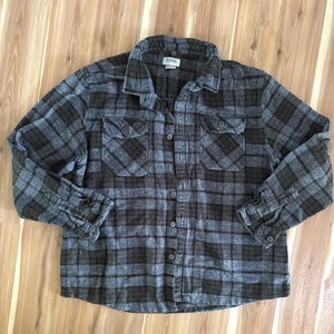 May include: A long-sleeve button-down shirt featuring a dark gray and brown plaid pattern. The shirt includes a standard collar, two chest pockets with buttoned flaps, and buttoned cuffs. A white label is visible at the inside of the collar.