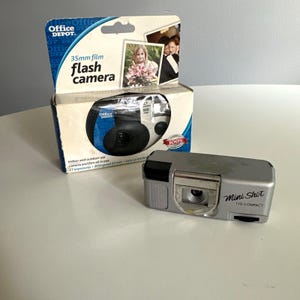 May include: A silver Mini Shot 110 compact camera with a black shutter button and a rectangular body. Behind it sits an Office Depot 35mm film flash camera box featuring a blue and white design with a photograph of a person holding flowers.