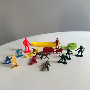May include: A collection of colorful plastic toy figures, including cowboys, Native Americans, horses, a covered wagon, and a canoe. The figures are in various poses and colors, creating a playful scene.