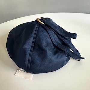 May include: A navy blue hat with a gold-colored metal ring and attached straps. The hat has a rounded shape and a tag attached to it. The fabric has a subtle sheen.