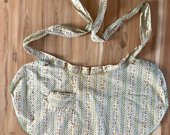 May include: A yellow and white floral print apron with a green scalloped edge and a single pocket. The apron has two long ties that can be tied around the waist.