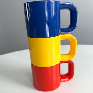 May include: Three stacked mugs in primary colors: blue, yellow, and red. Each mug has a rectangular handle. The mugs are arranged vertically, with the red mug at the bottom, the yellow in the middle, and the blue on top.