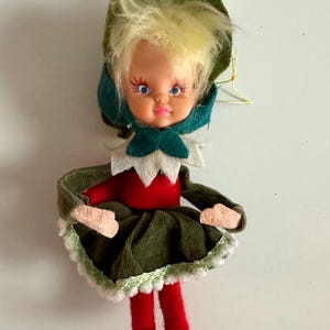 May include: A vintage doll ornament with a plastic face, blonde hair, and blue eyes. The doll wears a green hat, a red top, a green skirt, and red tights. The skirt is trimmed with white lace. A festive holiday decoration.