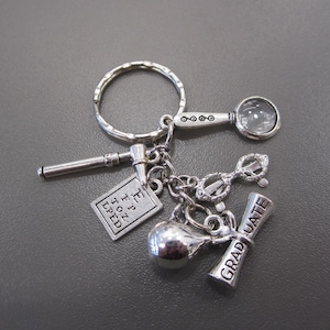 May include: A silver-toned metal keyring featuring multiple charms including a magnifying glass, a reflex hammer, a square eye chart with the letters E, FP, TOZ, LPED, a pair of glasses, a small round sphere, and a diploma scroll labelled GRADUATE.