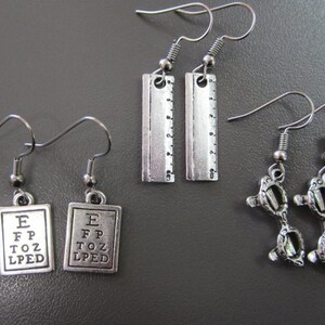 May include: Three pairs of silver dangle earrings. One pair features a silver eye chart with the letters E, FP, TOZ, and LPED. Another pair features a silver ruler. The third pair features a pair of silver glasses.