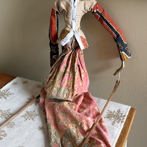 May include: An Indonesian Wayang Golek puppet. The puppet features a painted yellow face, red lips, and a detailed headdress. It wears a striped shirt, patterned fabric skirt, and has articulated arms. The puppet is held by wooden rods.