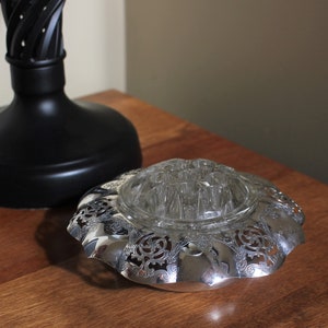 May include: A silver-plated floral arrangement holder with a clear glass insert. The holder has a decorative, ornate design with a scalloped edge.
