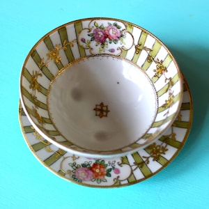May include: A white porcelain teacup and saucer set with a green and gold striped pattern and floral designs. The cup and saucer have a gold trim.
