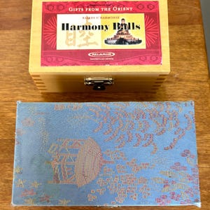 May include: A wooden box with a blue fabric lining. The box has a brass latch and a label that reads "Gifts from the Orient" and "Harmony Balls". The label also features a picture of a pagoda and a Buddha statue.