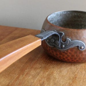 May include: Copper saucepan with a wooden handle. The hammered copper pot has a silver bracket attaching the handle. The handle is light brown and curved. The pot is on a wooden surface.