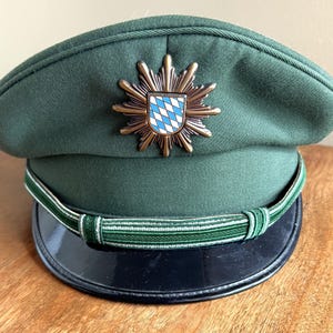May include: A dark green hat with a black brim and a decorative emblem. The emblem features a gold-coloured starburst design around a shield with a blue and white diamond pattern. A green and white striped band encircles the hat.