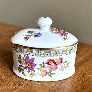 May include: A white ceramic sugar bowl with a lid, featuring a floral design in purple, pink, and yellow. The bowl has a gold rim and a decorative band with blue and yellow accents. The lid has a small, round handle.