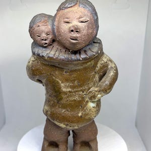 May include: A ceramic sculpture of two people, one carrying the other on their back. The sculpture is brown and white with a textured surface. The people have simple facial features and are wearing traditional clothing.