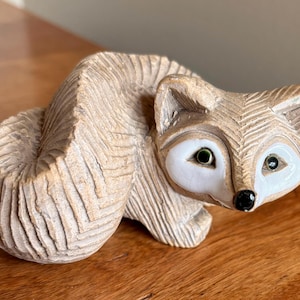 May include: A carved tan fox figurine with a textured, grooved body and a curled tail. The fox has smooth, white, glazed eye patches, small dark green eyes, and a glossy black nose. The sculpture features intricate, repeating chevron patterns.