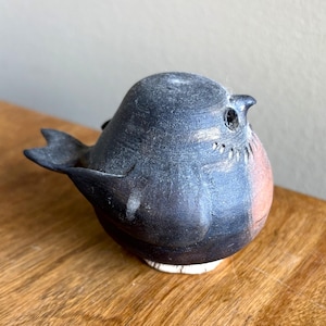 May include: A ceramic bird figurine with a rounded body and a small tail. The bird is primarily dark gray with reddish-brown and white accents. The figurine is sitting on a wooden surface.