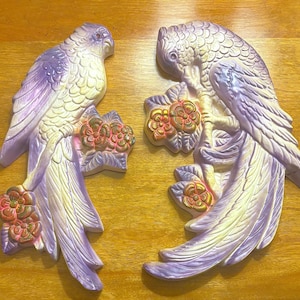May include: Two decorative parrot wall hangings. The parrots are in a light purple, cream, and gold color scheme. Each parrot is perched on a branch with flowers. The parrots have detailed feather and floral designs.
