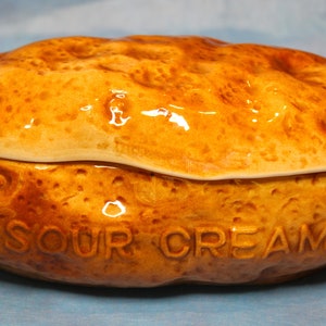 Peut inclure: Un plat à crème sure en céramique en forme de pomme de terre cuite. Le plat est brun avec une finition brillante et porte l'inscription "SOUR CREAM" en relief sur le côté. Le couvercle s'adapte parfaitement.
