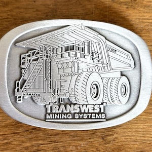 May include: An oval-shaped silver-toned metal belt buckle featuring an embossed graphic of a Komatsu 930E mining haul truck. The text "TRANSWEST MINING SYSTEMS" is engraved in a bold, sans-serif font at the bottom of the buckle design.