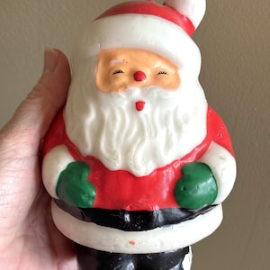 May include: A vintage Santa Claus candle. The candle features a red and white hat, a white beard, and a red suit with green gloves. The face is peach-colored with closed eyes and a red nose. The bottom is black.