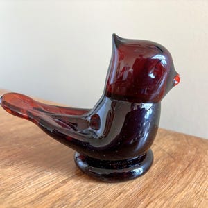May include: A dark red glass bird ornament with a rounded base and a long tail. The bird has a small red beak and a smooth, glossy finish. The ornament is on a wooden surface.