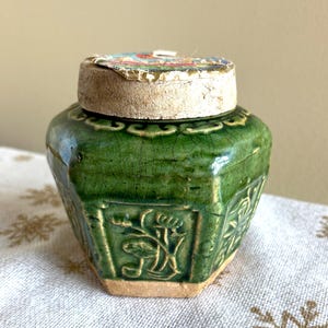 May include: A green ceramic jar with a lid. The jar has a hexagonal shape with embossed floral designs. The lid is made of a light-colored material with a colorful, patterned top. The jar has a crackled glaze finish.