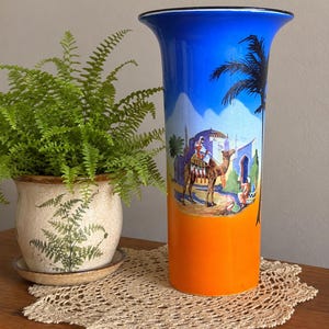 May include: A tall cylindrical vase featuring a vibrant gradient from bright blue at the top to orange at the base. The surface displays a detailed illustration of a camel and a person in a desert setting, accented by a black palm tree silhouette.