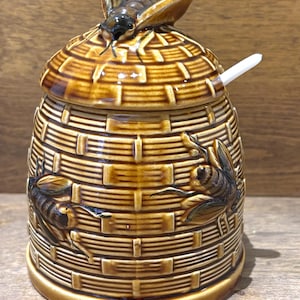 May include: A brown ceramic honey pot shaped like a beehive with a woven texture. The lid features a sculpted bee on top. Two additional bees are moulded onto the sides of the pot. A small white spoon is inserted into a notch on the side of the lid.