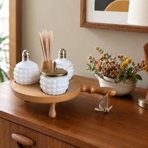 May include: A wooden serving tray holds three white, diamond-patterned glass jars with gold-colored lids and a wooden toothpick holder. The tray has a handle and a single wooden leg. A small floral arrangement and a lamp are in the background.