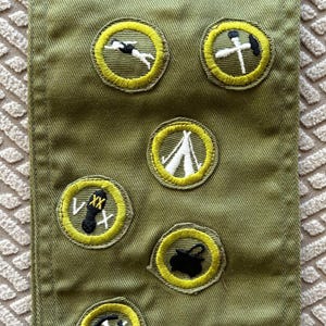 May include: A green fabric sash with several embroidered patches. The patches are yellow with black and white details. Some of the patches include a heart, a tent, a fish, and a pair of crossed axes.