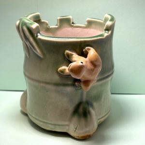 May include: A green ceramic planter with a frog design. The planter has a textured surface and a rounded base. The frog is perched on the side of the planter and has a light brown color.