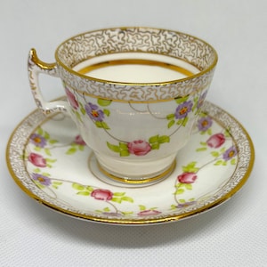 May include: A white teacup and saucer with a floral pattern. The cup and saucer have a gold trim and are decorated with pink and purple flowers.