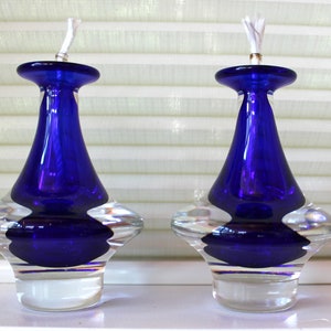 May include: Two cobalt blue glass oil lamps with clear glass bases. The lamps have a round, wide base and a narrow neck with a wick.