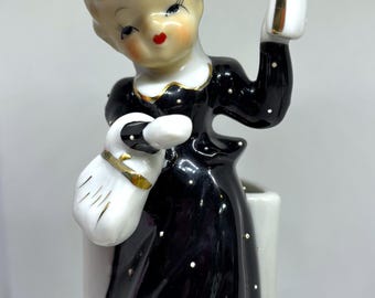 May include: Vintage ceramic figurine of a woman in a black dress with white polka dots, holding a white purse and a white object with gold trim. The figurine has a black hat with a pink flower and is a decorative item.