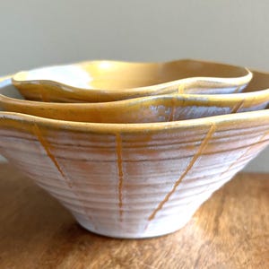 May include: A set of three ceramic bowls in a yellow and white glaze. The bowls are nested inside each other and have a wavy, organic shape.