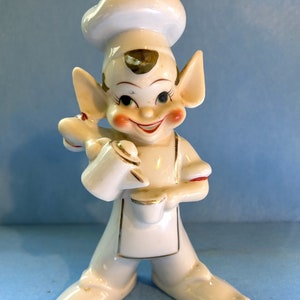 Peut inclure: Une figurine en céramique blanche d'un lutin souriant portant un chapeau de chef et tenant un bol à mélanger. Le lutin a les yeux verts, les joues roses et un tablier à bordure dorée.