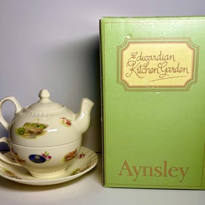 May include: A cream-colored teacup, saucer, and teapot set with a lid, featuring a floral and fruit design. The set is next to a green box with the words "Edwardian Kitchen Garden" and "Aynsley" printed on it.