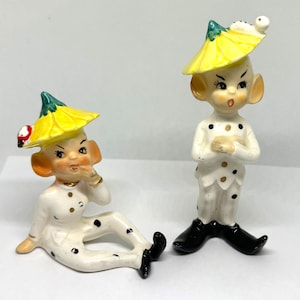 May include: Two ceramic figurines of elves wearing yellow and green hats with black polka dots. The elf on the left is sitting with its hand on its chin, while the elf on the right is standing with its arms crossed.