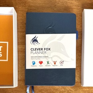May include: An overhead shot of three items: a white box containing an orange booklet, a navy blue planner with a white label, and a white box with a logo. The booklet reads "HOW IT WORKS" and "WEEKLY PLANNER". The planner is labeled "CLEVER FOX PLANNER".