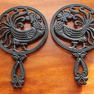 May include: Two black cast iron rooster hot pads with intricate designs. The roosters are facing each other with their tails spread out.