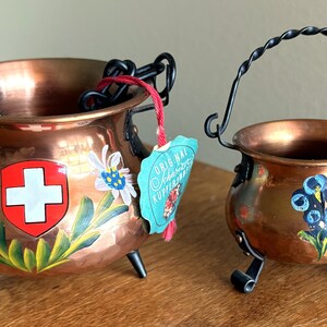 Peut inclure: Deux petites marmites en cuivre avec des motifs floraux peints et des poignées en métal noir. La plus grande marmite a un drapeau suisse peint dessus et une étiquette qui indique "Original Schweizer Kupferbirn". La plus petite marmite a une fleur bleue peinte dessus et le texte "St. Gall" sur le côté.