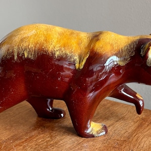 May include: A ceramic bear figurine featuring a glossy finish with a deep mahogany brown body and a contrasting golden yellow glaze across the back, ears, and paws. The bear is depicted in a walking stance with its head lowered and one front paw raised.