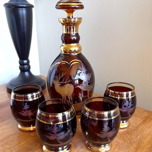 Peut inclure: Un service à décanter en verre ambré vintage avec un motif de cerf et de feuilles. L'ensemble comprend une carafe avec un bouchon et quatre verres assortis, tous avec des bordures dorées. Les verres et la carafe sont gravés d'un motif inspiré de la nature.